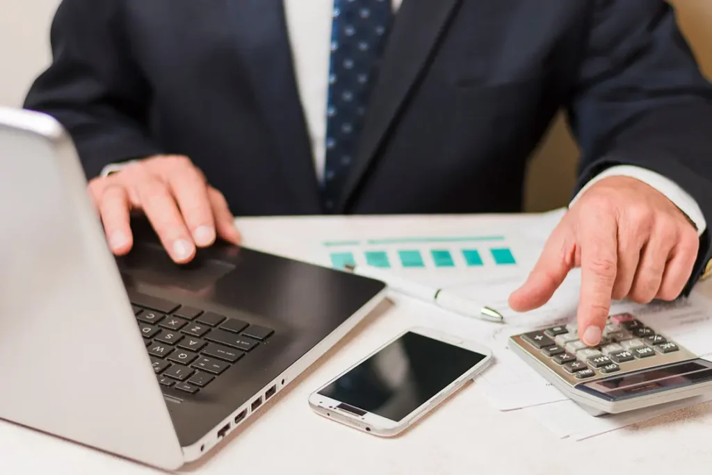 Homem de terno e gravata em uma mesa de trabalho operando um computador com a mão direita e uma calculadora com a mão esquerda. Sobre a mesa, um celular e papéis com gráficos.