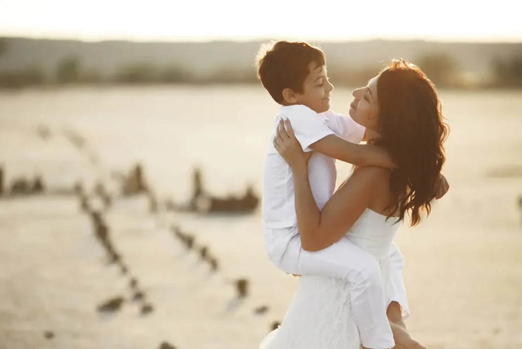 Mãe jovem, de cabelo preto e cacheado, vestida de branco, segurando menino, também todo vestido de branco, no colo. Os dois se olham sorrindo.