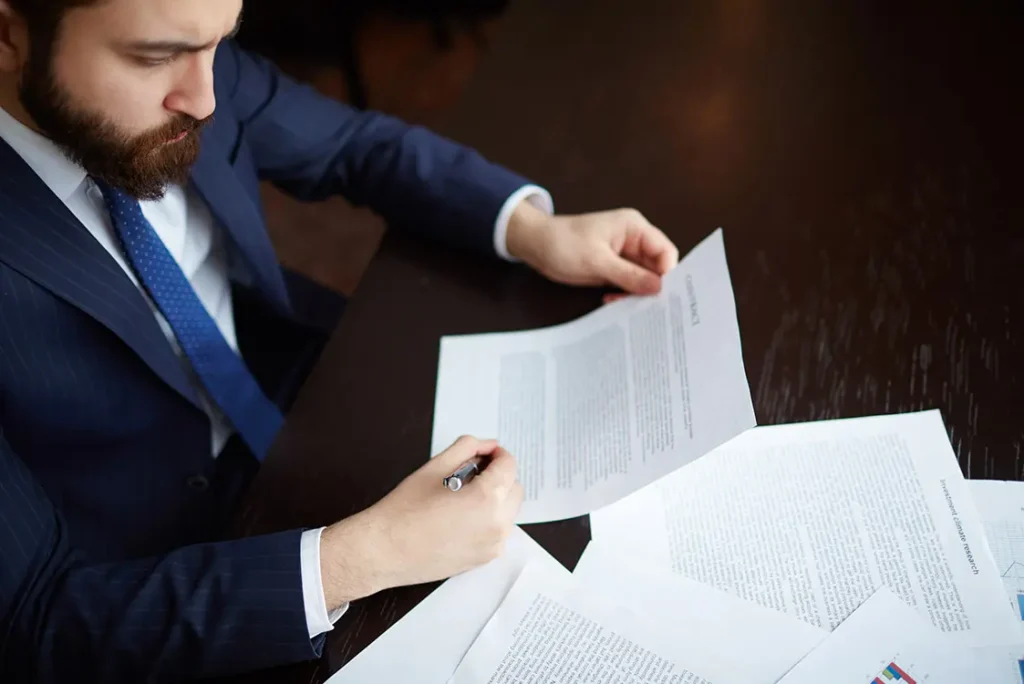 Homem barbudo de terno azul sentado à mesa com um documento na mão e uma série de outros documentos à sua frente.