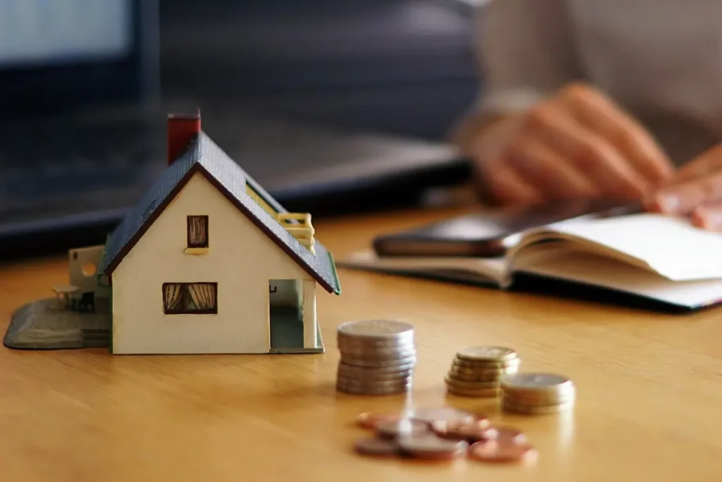 Mesa com miniatura de uma casa, notas de dinheiro amassada e moedas, sobre ela
