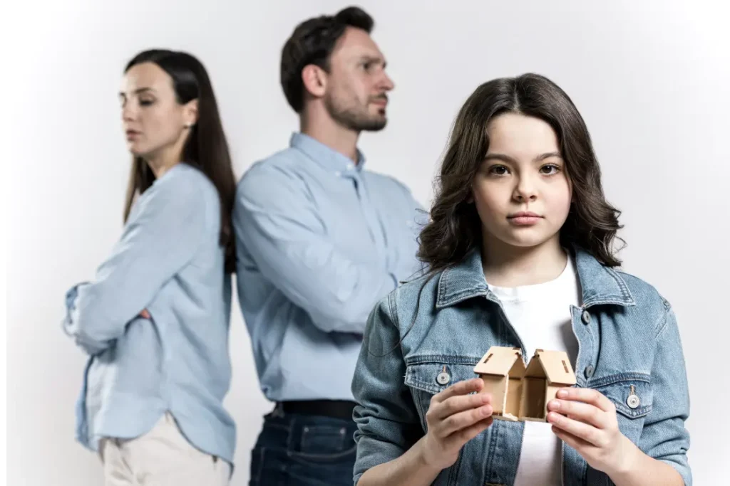 menina com uma casa de brinquedo, partida, nas mãos. Ao fundo, seus pais de costas um para o outro