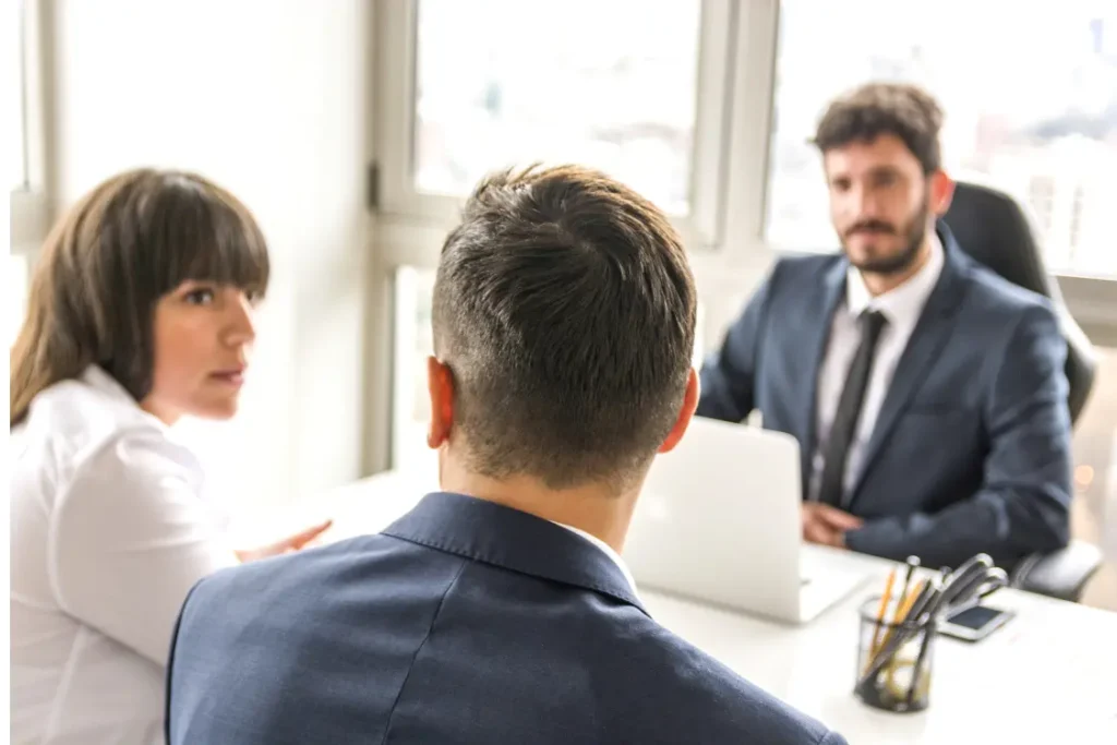 Mulher acompanhada de um homem sentada à frente de uma mesa de escritório. Do outro lado, outro homem, ouve o que eles dizem
