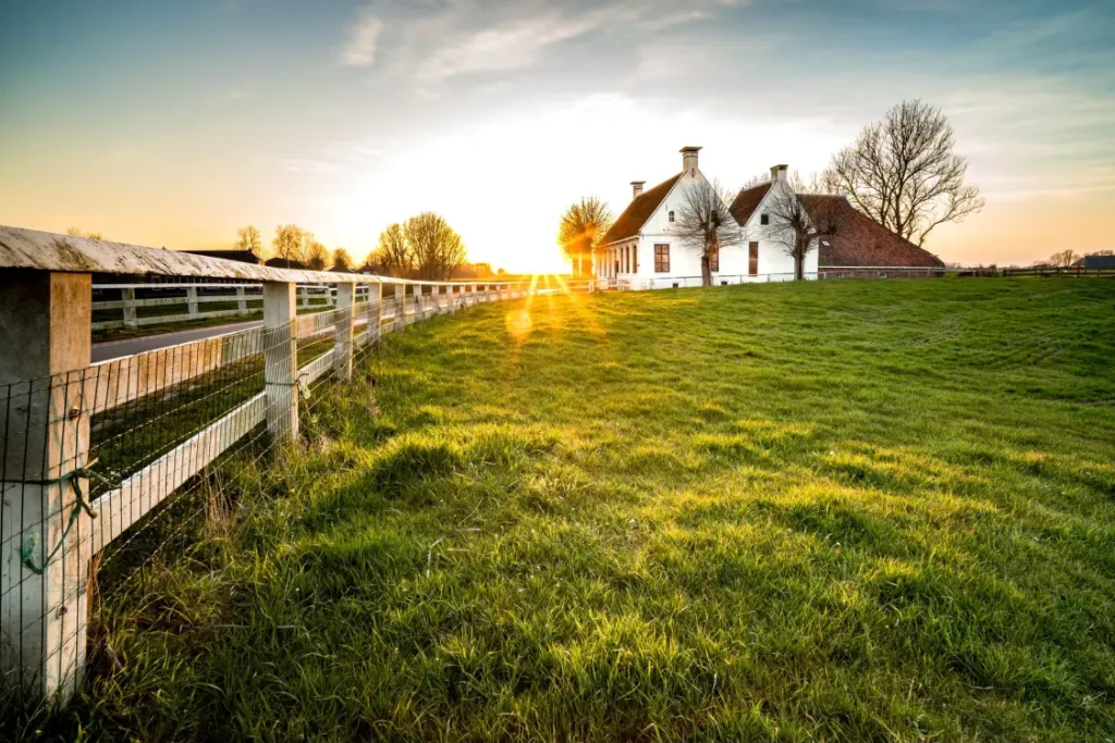 por do sol em uma propriedade rural, com um pasto verde e cerca de madeira. Ao fundo, uma casa de campo